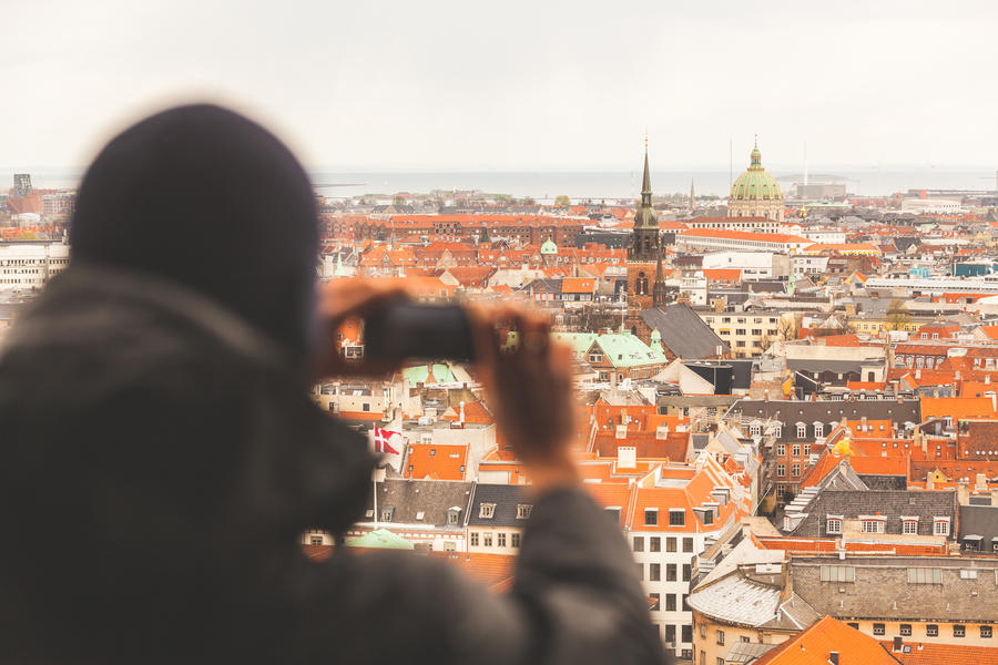Aerial photo of Copenhagen on a cloudy day with a tourist taking a picture with his smart phone. He is out of focus, on the left of the frame. A lot of red roofs and bell towers are visible all around