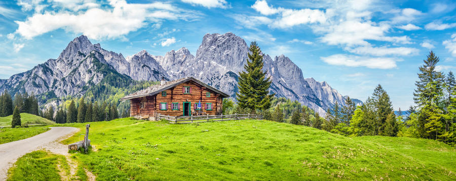 Panoramic view of beautiful mountain scenery in the Alps with traditional rural mountain chalet and fresh green meadows on a sunny day with blue sky and clouds in spring