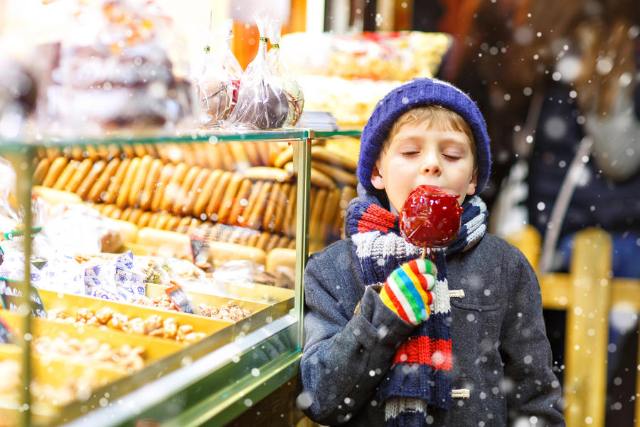 Little cute kid boy near sweet stand with gingerbread and nuts. Happy child eating on apple covered with red sugar. Traditional sweet on German Christmas market.