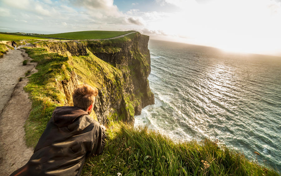 Young man laying down on the edge of the cliffs on the Coastal footpath walk, Cliffs of Moher, huge sea cliffs located at the southwestern edge of the Burren region in County Clare, Ireland.