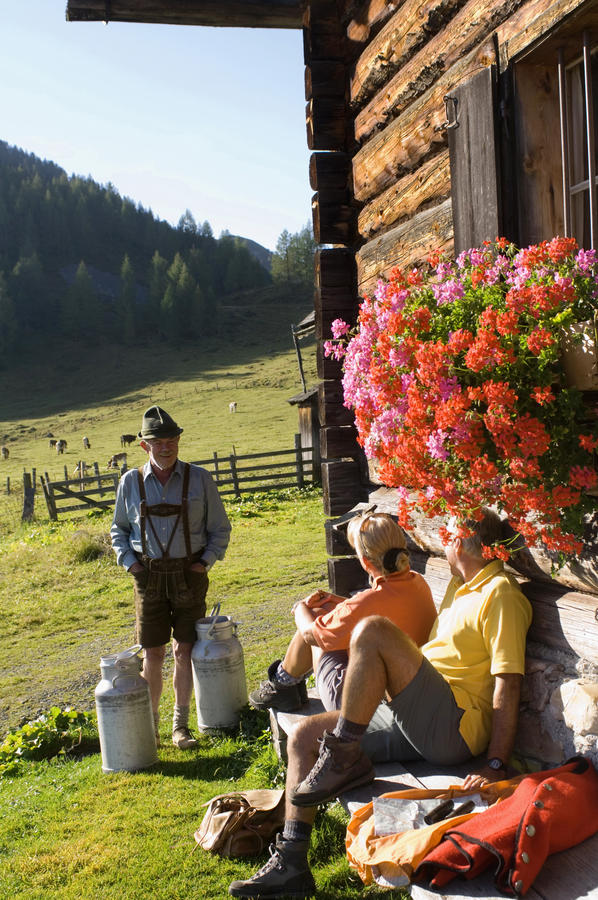 Mature couple sitting in front of alpine hut while farmer carrying milk cans