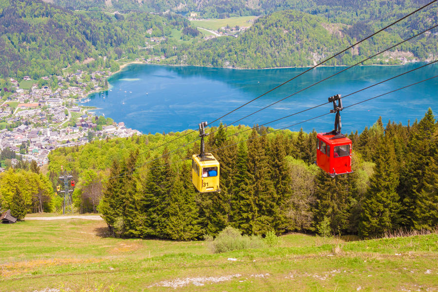 Yellow and red gondolas of Zwoelferhorn Seilbahn (cable way) travel up and down alpine peak with a view of village St.Gilgen and Wolfgangsee lake, Salzburg land, Austria