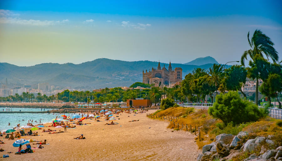 View of the beach of Palma de Mallorca with people lying on sand and the gorgeous cathedral building visible in background. Palma-de-Mallorca, Balearic islands, Spain.