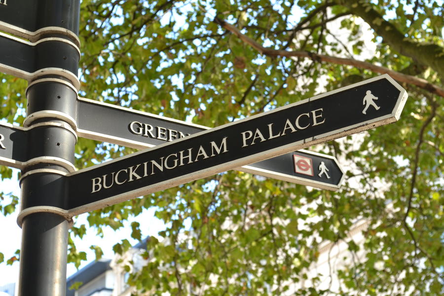 London Buckingham Palace direction signpost near Hyde Park Corner with green tree branches in the background.
Direction signpost to British Queen London residence The Buckingham Palace.