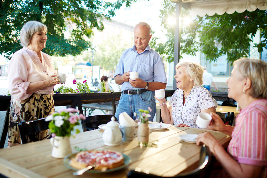 Cheerful senior friends celebrating holiday in outdoor cafe while one of them making toast, teapot and tasty pie standing on wooden table