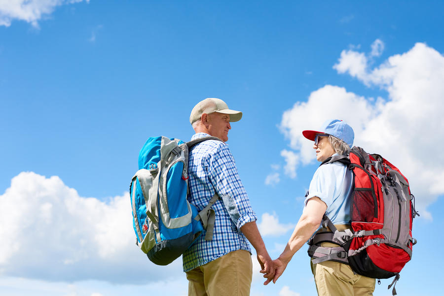 Back view portrait of active senior couple travelling on hiking trip, both wearing backpacks, standing holding hands against clear blue sky, copy space