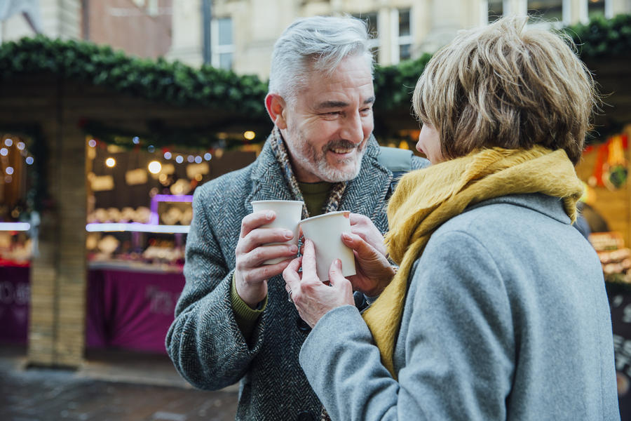 Mature couple are drinking hot drinks in a town christmas market. They are toasting the disposable cups.