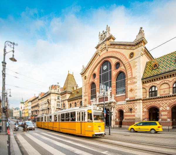 Central Market Hall in Budapest city, Hungary, Europe. Pedestrian crossing and yellow tram in foreground, old building and blue sky in background.